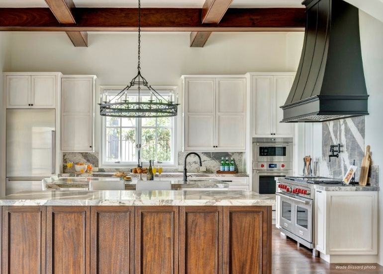 Raw Urth Montrose Modern kitchen with wooden beams, white cabinets, marble countertops, large island with dark wood paneling, stainless appliances, natural light over the sink, and a Raw Urth Montrose black range hood as a striking focal point.