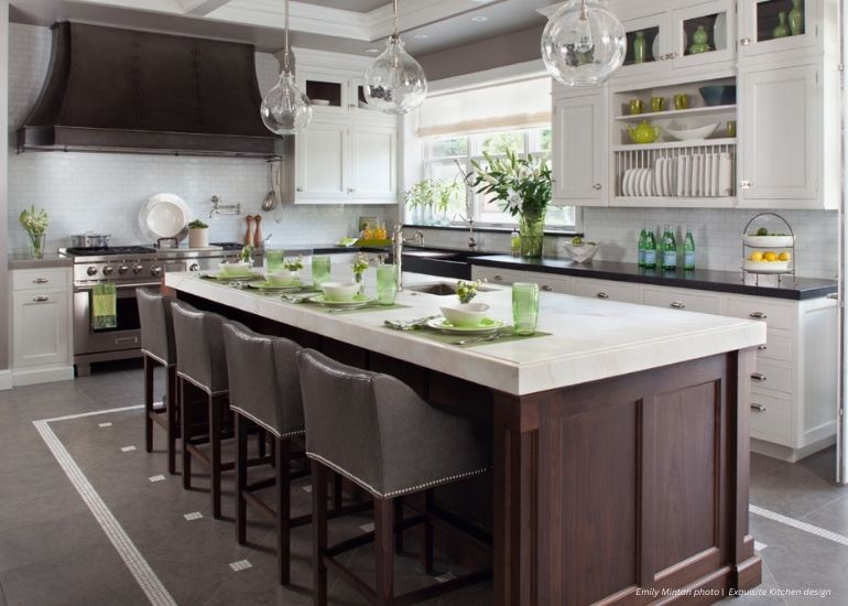 Raw Urth Winter Park Modern kitchen featuring a Raw Urth Winter Park island with a dark wood base and white countertop. Four gray upholstered barstools, green-tinted glassware, white cabinets, and stainless appliances complete the look.