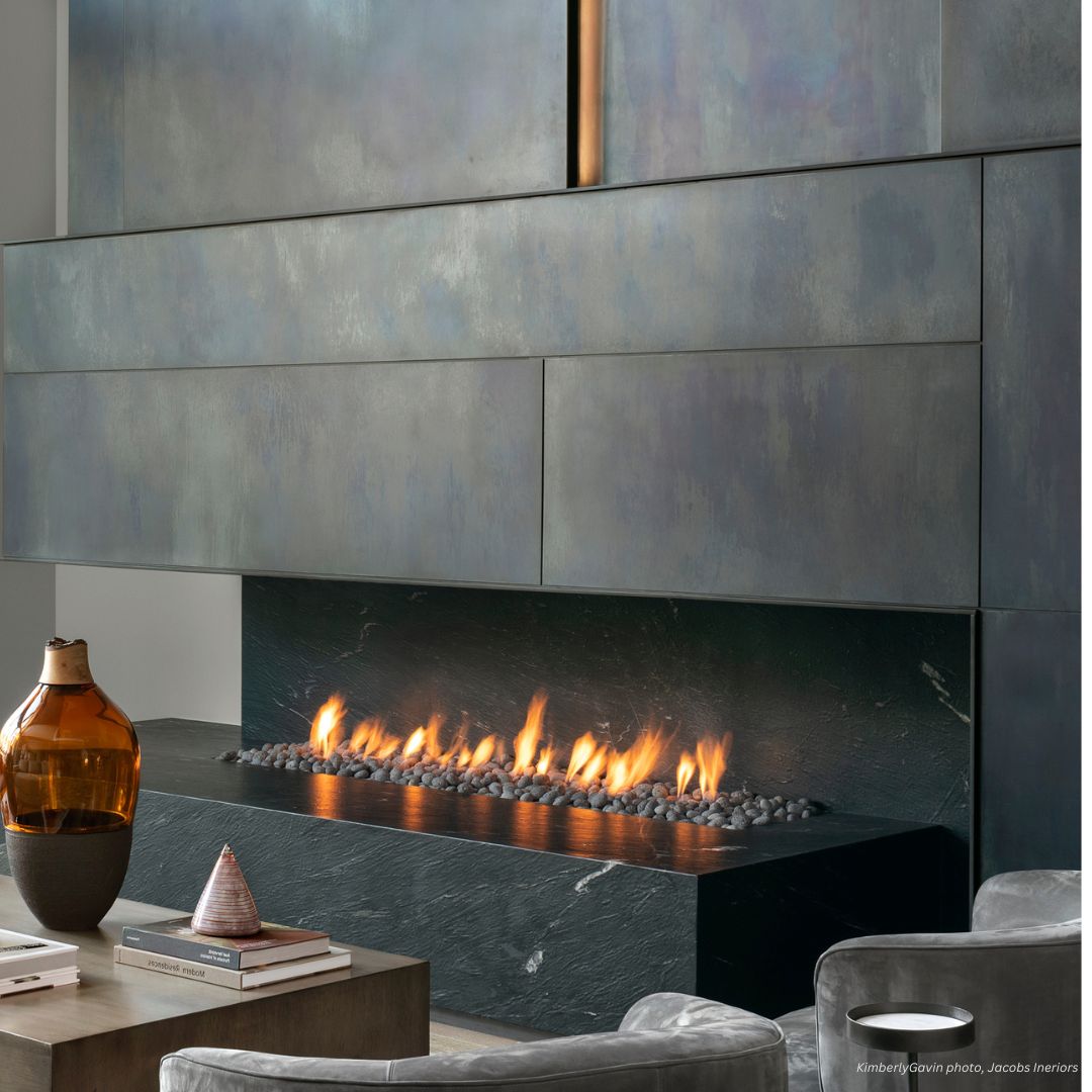 A modern living room featuring a fireplace and mantel from Portfolio_Raw Urth Designs set in dark stone, with minimalist gray cabinetry above and contemporary furniture accented by a glass vase and decor on the coffee table.