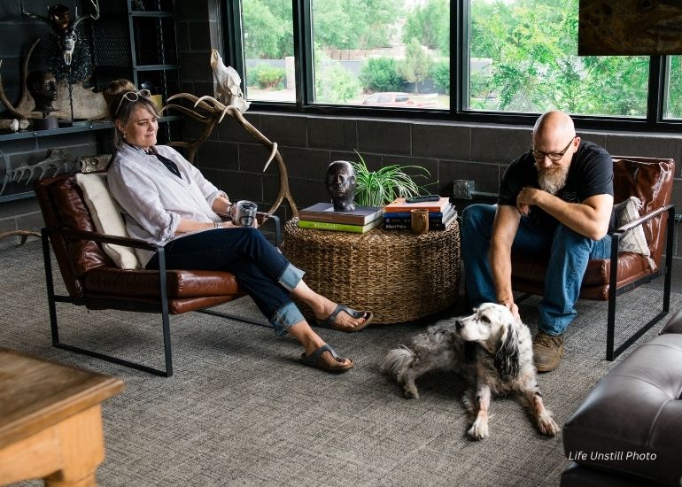 In a bright, modern room, a man pets a dog lying between him and a woman as they relax. The ru-amysteffandog-tablet sits on a wicker table nearby, surrounded by books and decorations beneath large windows.