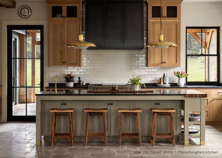 A modern kitchen with ru-aspen light wood cabinets, a large island and three wooden stools, features a white subway tile backsplash, black range hood, two gold pendant lights, and glass doors allowing natural light.