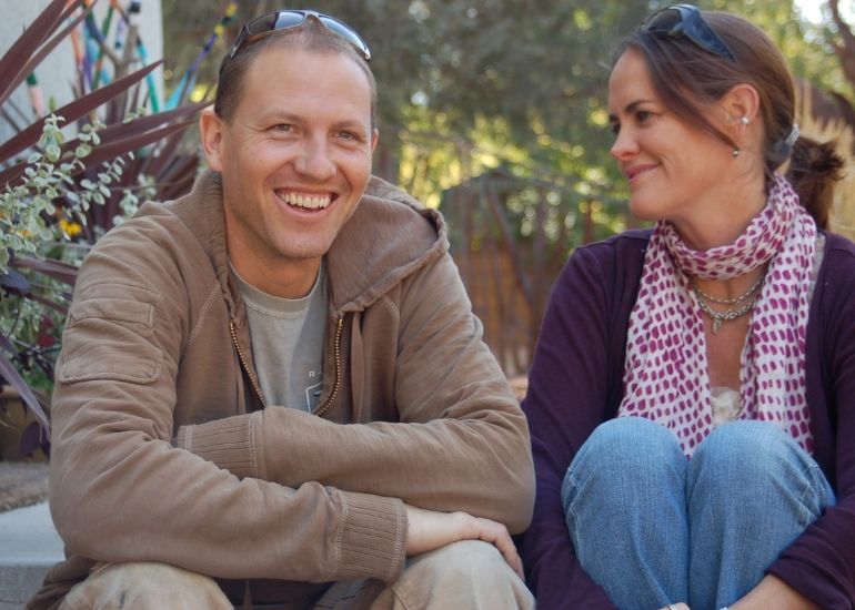 A man, wearing ru-chapter1—a brown jacket with sunglasses on his head—sits outdoors smiling at a woman in a scarf and necklaces. They enjoy each other's company among trees and garden plants.