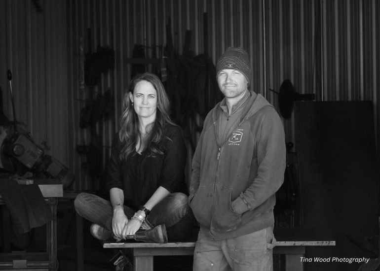 A woman sits cross-legged on a workbench next to a standing man, both facing the camera in a workshop with metal tools and equipment. This black and white image features the ru-chapter2 product.