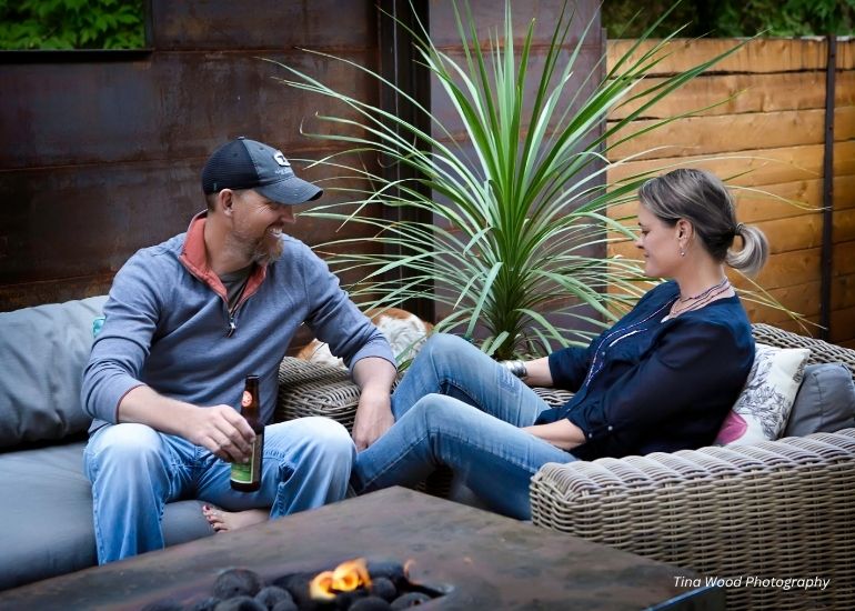 A man and woman smile and relax on the ru-chapter3 patio sofa by a fire pit. The man holds a beer as they sit casually dressed, with green plants and wooden fencing in the background.