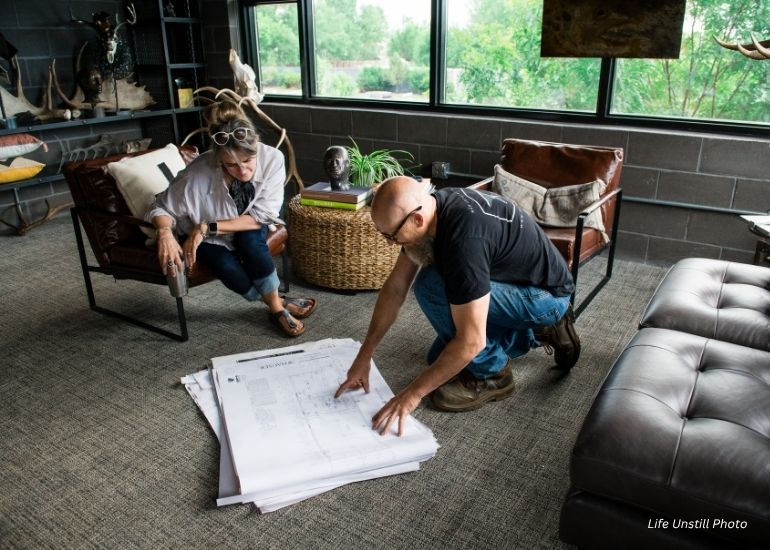 A man points at large blueprints on the floor as a woman watches from an armchair in a modern office with large windows, featuring the ru-chapter4 product.