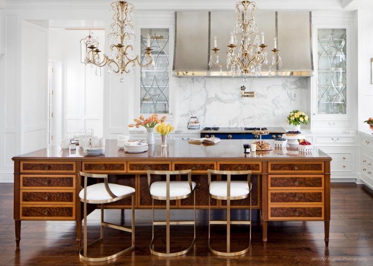The ru-contact kitchen features a large wooden island, three modern chairs, two chandeliers, white cabinetry, a marble backsplash, stainless steel range, and counters adorned with fresh flowers, dishes, and pastries.