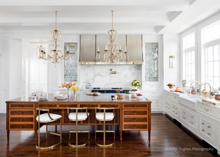 ru-contact Bright, elegant kitchen featuring the ru-contact large wooden island, gold-accented chairs, two chandeliers, marble backsplash, stainless steel stove, white cabinetry, and expansive windows for natural light.