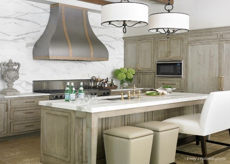ru-custom-bellvue The ru-custom-bellvue features an elegant kitchen with light wood cabinets, a marble backsplash, a spacious marble island topped with drinks and flowers, modern pendant lights overhead, plus upholstered stools and a white chair for seating.