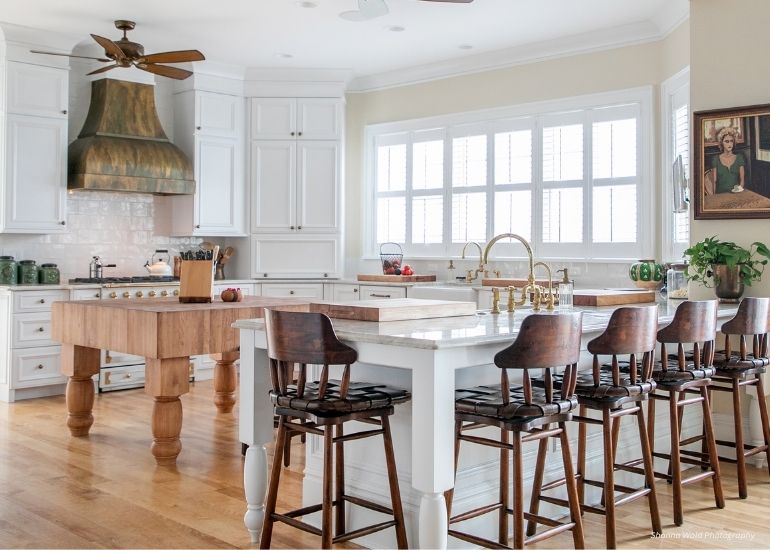 The ru-custom-bonnerpeak kitchen features white cabinets, a large marble island with four wooden bar stools, a butcher block table, brass fixtures, hardwood floors, a copper range hood, and sunlit shuttered windows.