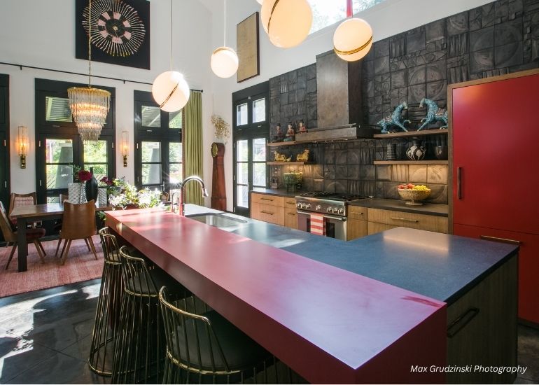 Modern kitchen with the ru-custom-boulder bold red island countertop, black backsplash, wooden cabinets, and gold bar stools; dining area with chandelier and large windows in the background.