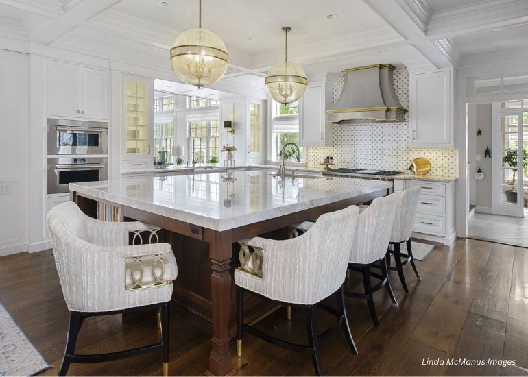 ru-custom-cherryhills Modern kitchen featuring the ru-custom-cherryhills, a large marble island with six upholstered chairs, pendant lights, stainless steel appliances, white cabinetry, wood floors, and sunlit open space through expansive windows.