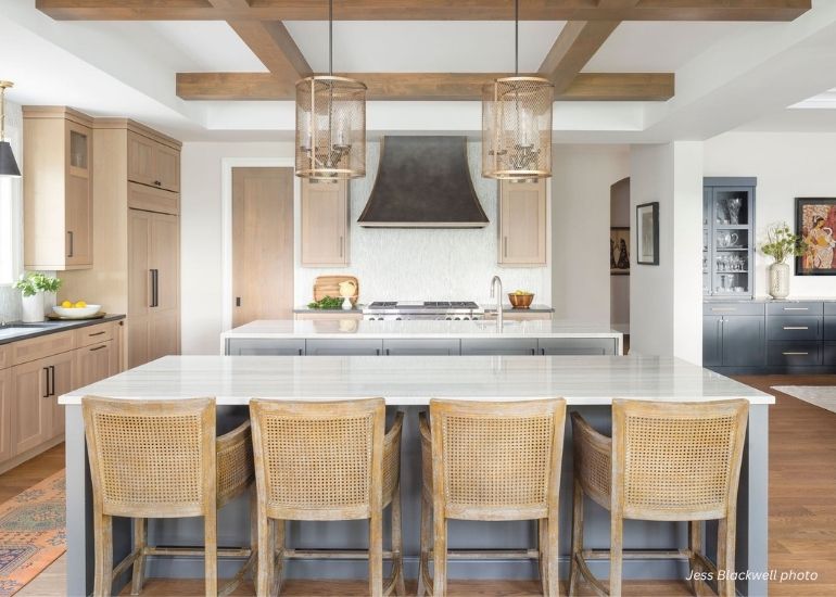 ru-custom-dovecreek The ru-custom-dovecreek kitchen features a spacious island with four wicker bar stools, light wood cabinets, a stainless steel stove with dark vent hood, wooden ceiling beams, and two box pendant lights.