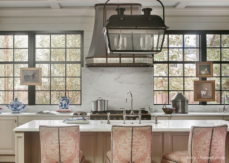 ru-custom-durango A bright kitchen with large windows showcases the ru-custom-durango range hood, a marble backsplash, white island with three pink-patterned chairs, and blue-and-white ceramics and art decorating the counters.