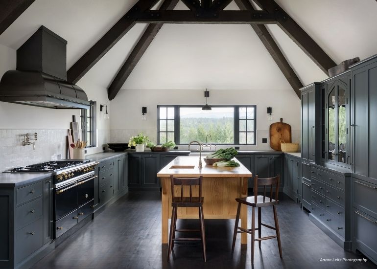 ru-custom-palisade A modern farmhouse kitchen featuring ru-custom-palisade dark cabinetry, a wood island with seating, exposed beams, large scenic windows, stainless steel appliances, and counters adorned with fresh produce and kitchenware.