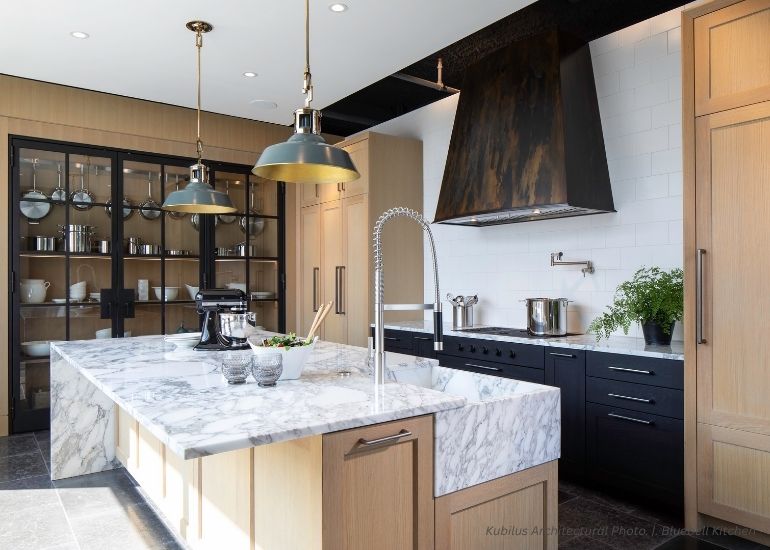 The ru-custom-tabernash features a marble island, wood cabinetry, black stove, large metal range hood, glass-fronted cabinets with dishes, pendant lights, tall faucet, plus fresh herbs and utensils on the counters.