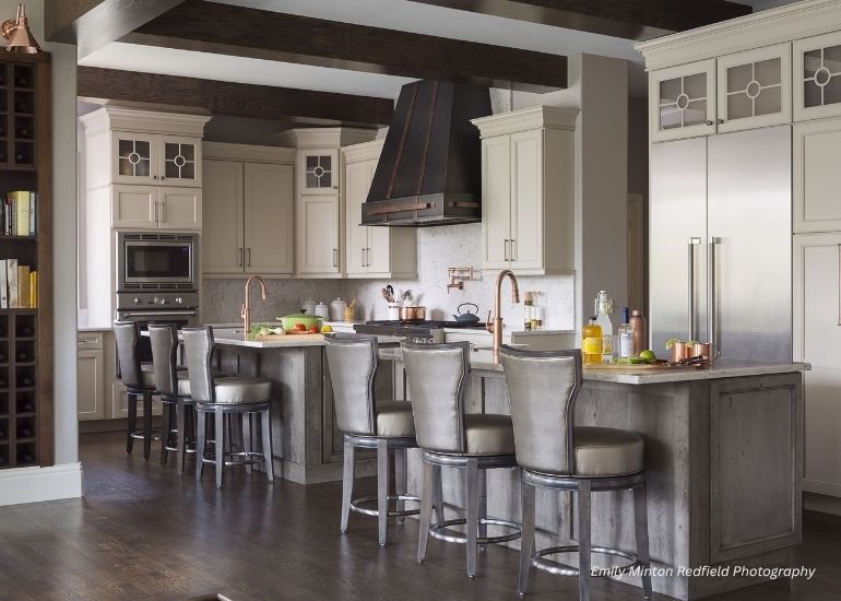 Modern kitchen with ru-custom-vail island, seating for four, stainless steel appliances, cream cabinetry, and copper accents. Fresh produce and dishware decorate the countertops. Dark wood floors enhance the stylish space.