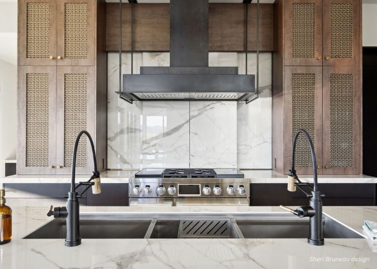 Modern kitchen featuring ru-custom-washpark: marble backsplash, stainless steel stove, dual sinks with black faucets, woven wood cabinets, and a large vent hood above the stove.