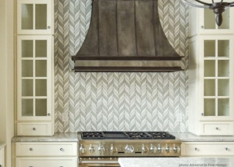 The kitchen includes a ru-faq-tablet stove with six burners, a dark range hood, glass-front cabinets flanking it, and a gray and white chevron tile backsplash.