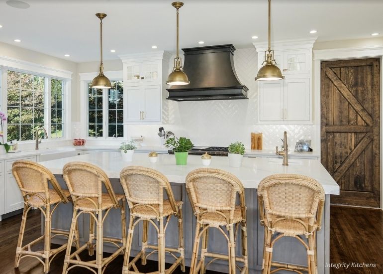 ru-montrose The ru-montrose kitchen features white cabinets, a spacious island with a white countertop, five wicker barstools, brass pendant lights, a black range hood, and a rustic wooden sliding door.