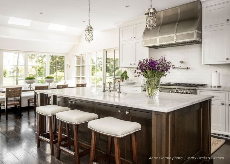 The ru-portfolio-custom features a bright, modern kitchen with a large dark wood island, white countertops, three upholstered stools, stainless steel appliances, white cabinets, and a vase of purple flowers. Sunlight floods in through large windows.