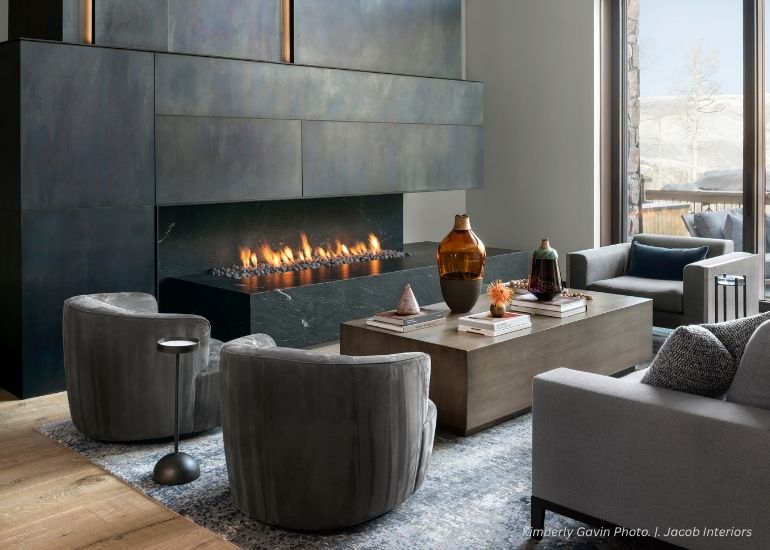 ru-portfolio-fireplace A modern living room features gray swivel chairs, a wooden coffee table with vases and books, and the ru-portfolio-fireplace—a sleek linear gas unit set in a dark metal wall. Large windows display a snowy outdoor view.