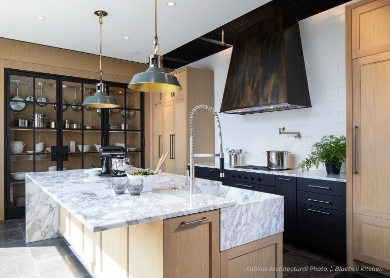 ru-portfolio-predesigned The ru-portfolio-predesigned kitchen features a marble island, wooden cabinets, black appliances, open display shelves, two green pendant lights over the island, and a large metal vent hood above the stove.