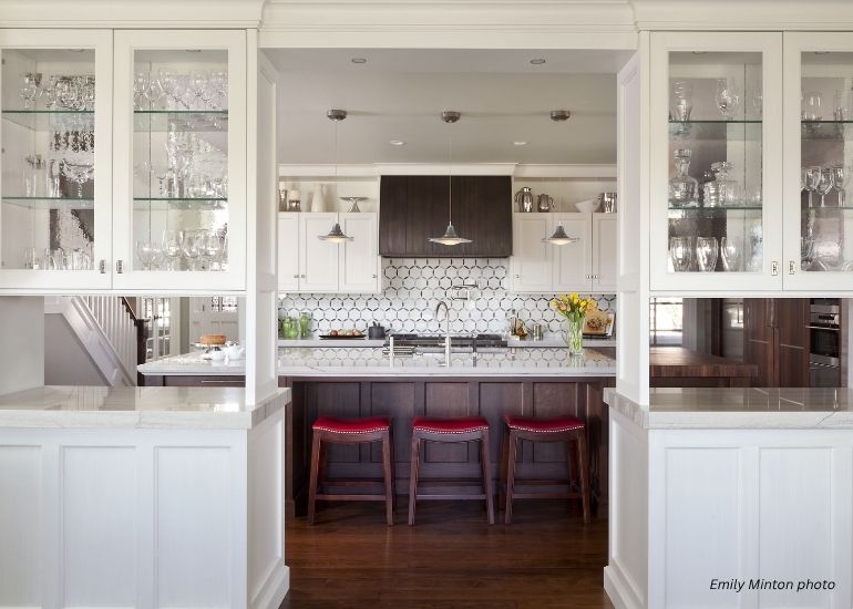 The ru-snowmass kitchen features white cabinets, glass display shelves, a hexagonal tile backsplash, stainless steel appliances, a central island with three red stools and pendant lights above. Yellow flowers brighten the island.