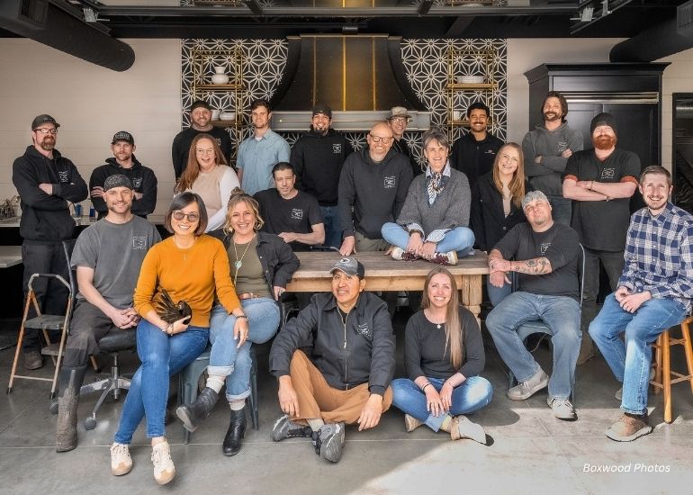 A group of 19 casually dressed people, likely a restaurant team, pose together—some sitting, some standing—in a modern industrial kitchen with the ru-team-tablet. Most are smiling for the photo.