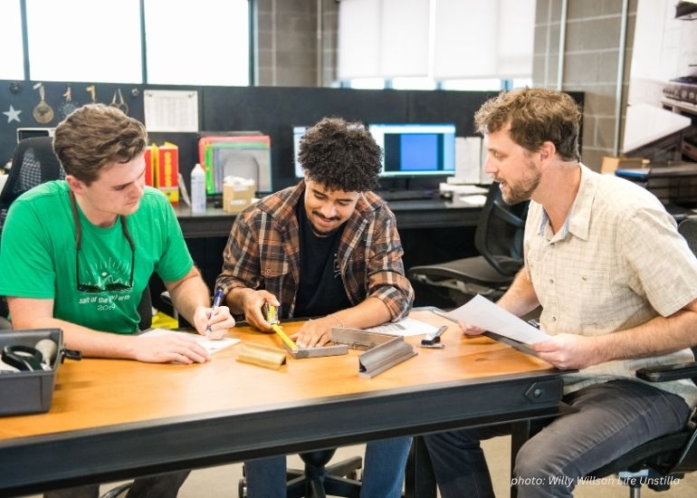 Three men collaborate at a workbench using ru-team to measure wooden pieces and take notes, with office supplies and computer monitors in the background.