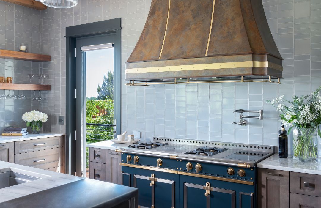 Modern kitchen features a La Cornue Oven with brass, Raw Urth Steel range hood in brass and patina steel, open wooden shelves, marble countertops, vases of white flowers, and a glass door overlooking greenery.