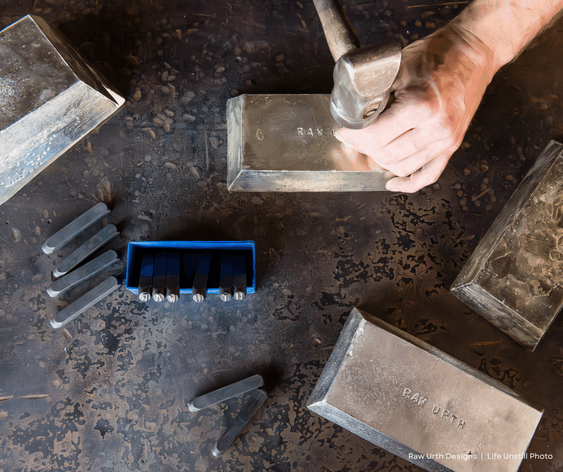 A person uses product 1 and a metal stamp to engrave text onto a silver bar; other stamped bars, loose stamps, and a blue stamp container are on a textured metal work surface.
