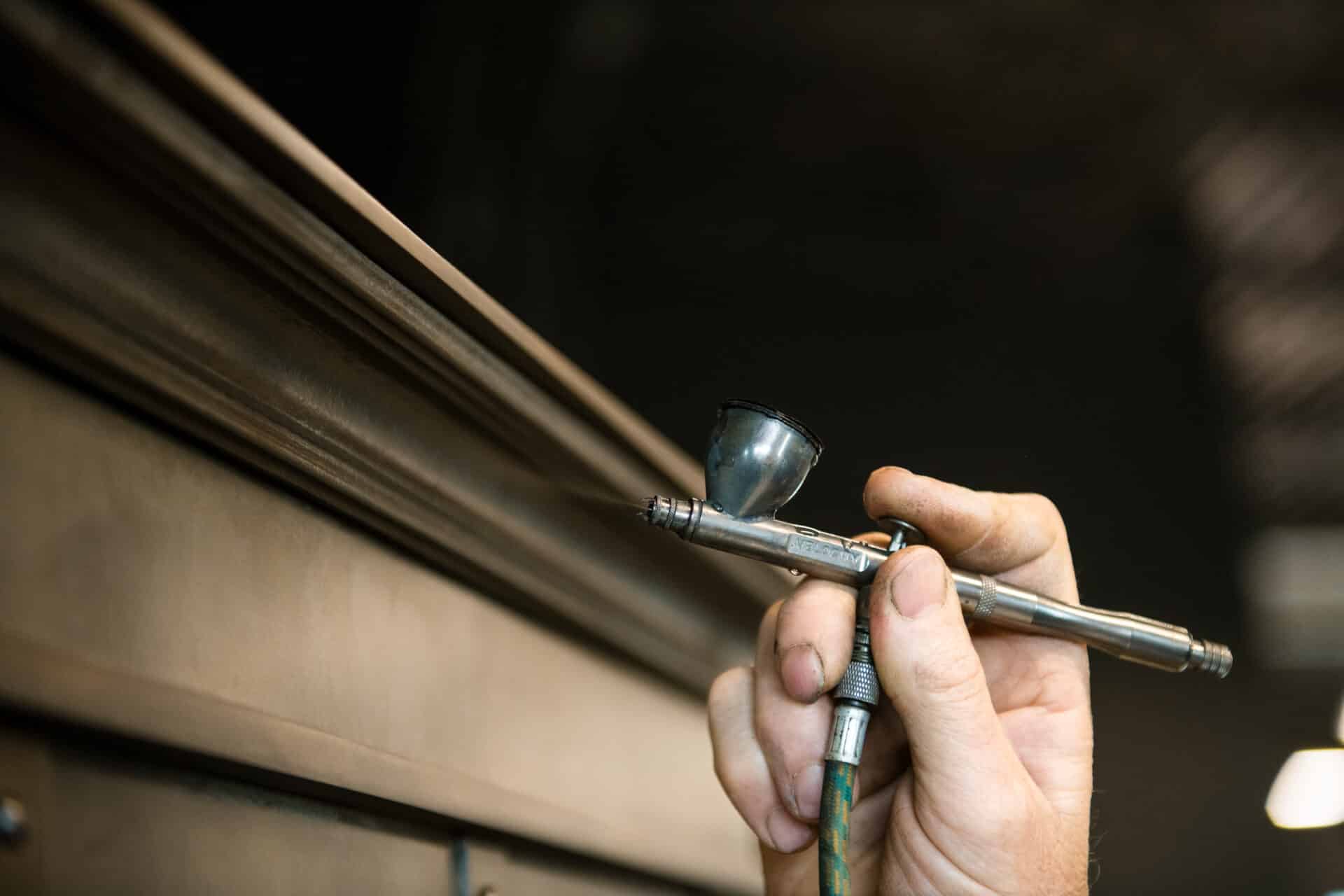 A close-up shows a hand using an airbrush tool to finish the edge of wood, highlighting www.lifeunstill.com. The background is blurred for focus on the process.