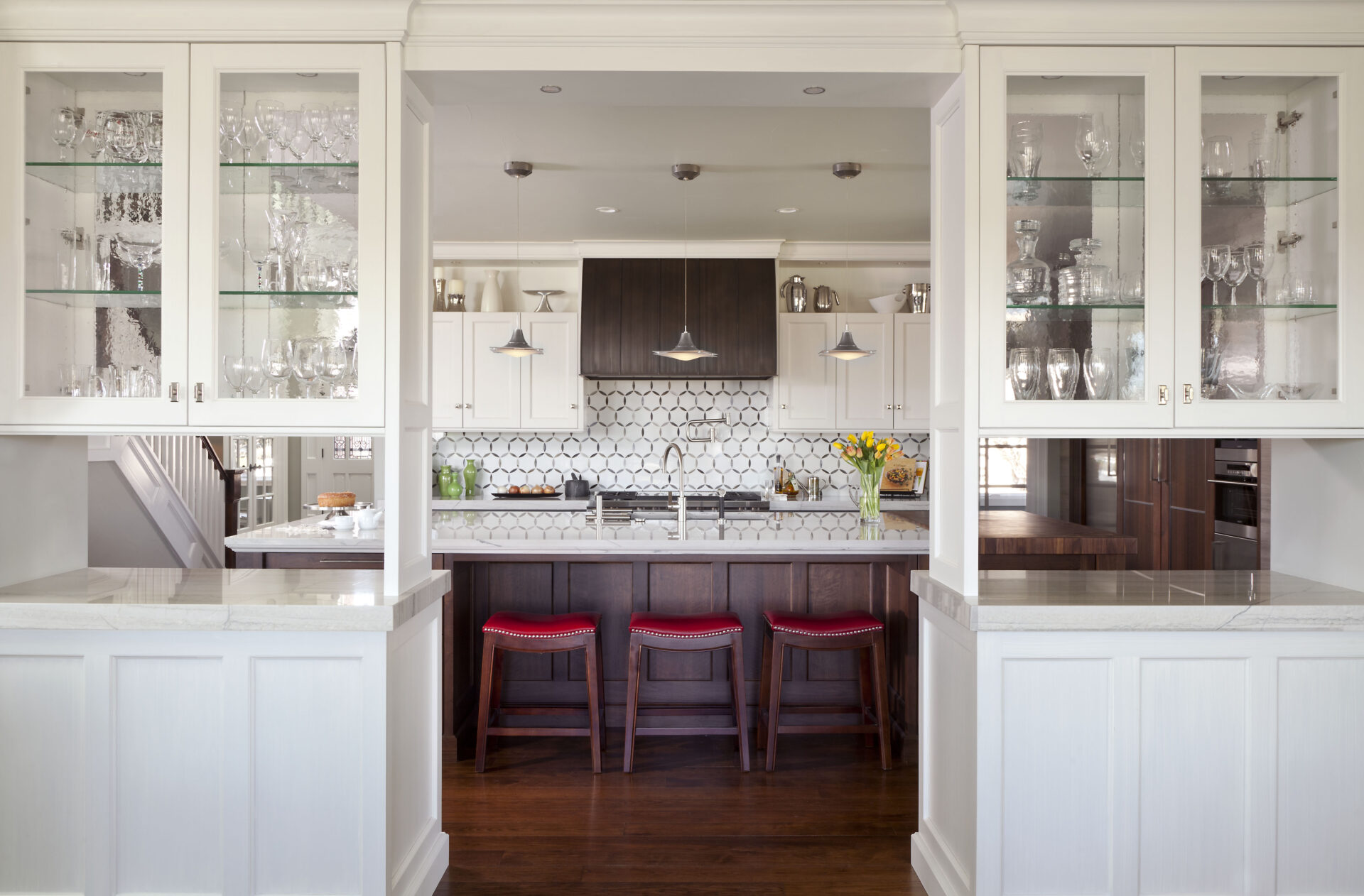 A modern kitchen featuring Raw Urth Designs Steel Range Hood Aspen Snowmass Brushed panels with standing ribs and rivets, white cabinetry, glass shelves, hexagonal tile backsplash, three wooden bar stools with red seats, and pendant lighting.