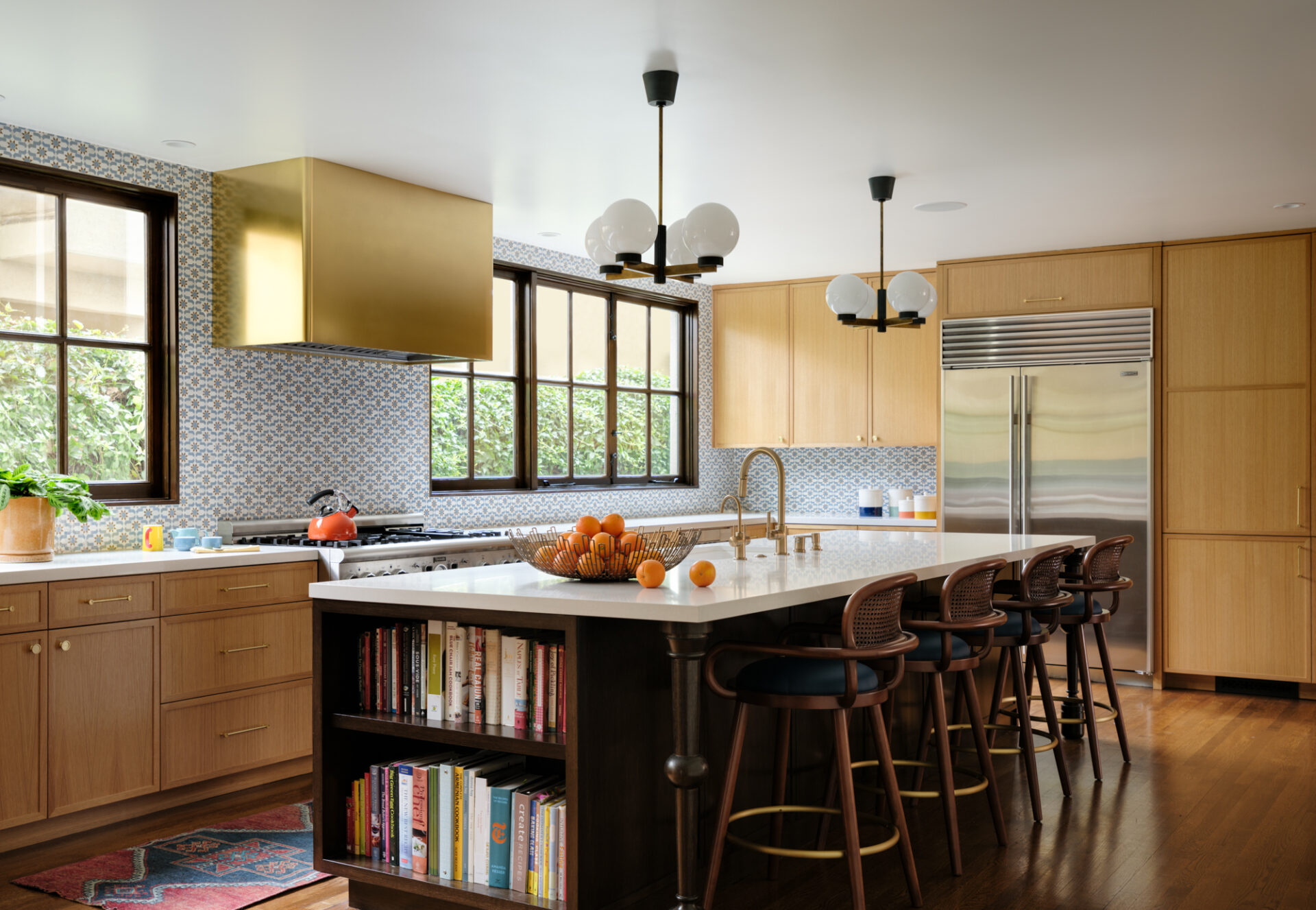 Modern kitchen with Brass Raw Urth Range Hood Broadmoor, white island with bar stools, built-in bookshelf, stainless appliances, globe pendants, windows, and a bowl of oranges on the counter.