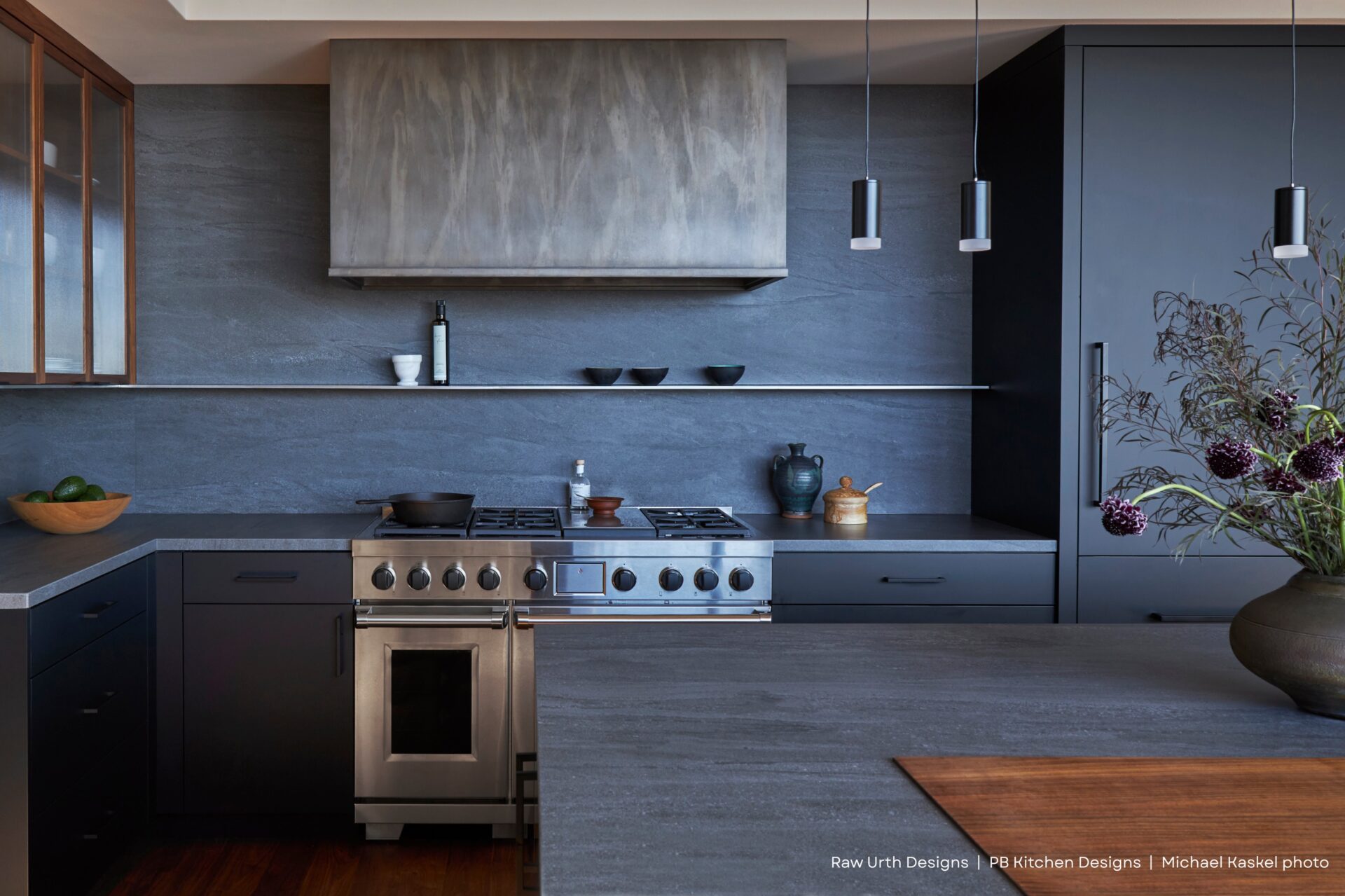 Modern kitchen featuring Raw Urth Designs by PB Kitchen Designs and Michael Kaskel—matte dark cabinets, stainless steel stove and hood, minimalist shelving with dishes, a large island topped with flowers, and warm wooden floors for a sleek look.