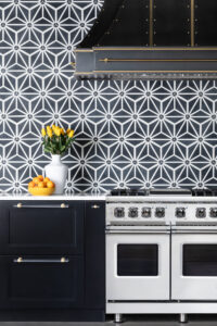 Modern kitchen with black and white geometric tile backsplash, black cabinetry with gold handles, a white vase of yellow tulips, a bowl of oranges, and a stainless steel stove featuring the Raw Urth Showroom Hood Montrose Double Stack with burnished brass trim.