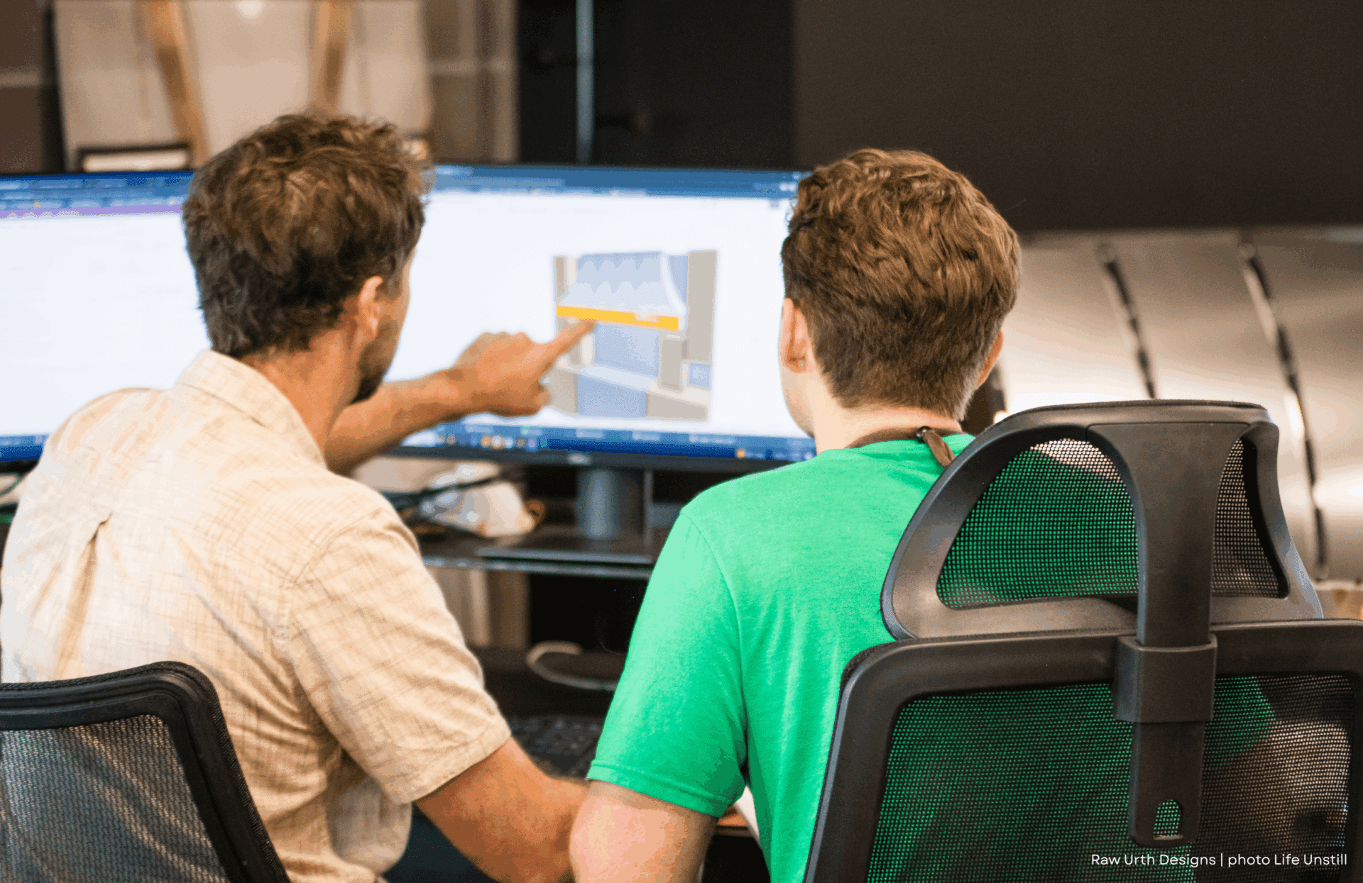 Two people sit at 2 desks in front of large computer monitors—one points at a screen showing a technical diagram or 3D model, while the other observes attentively.