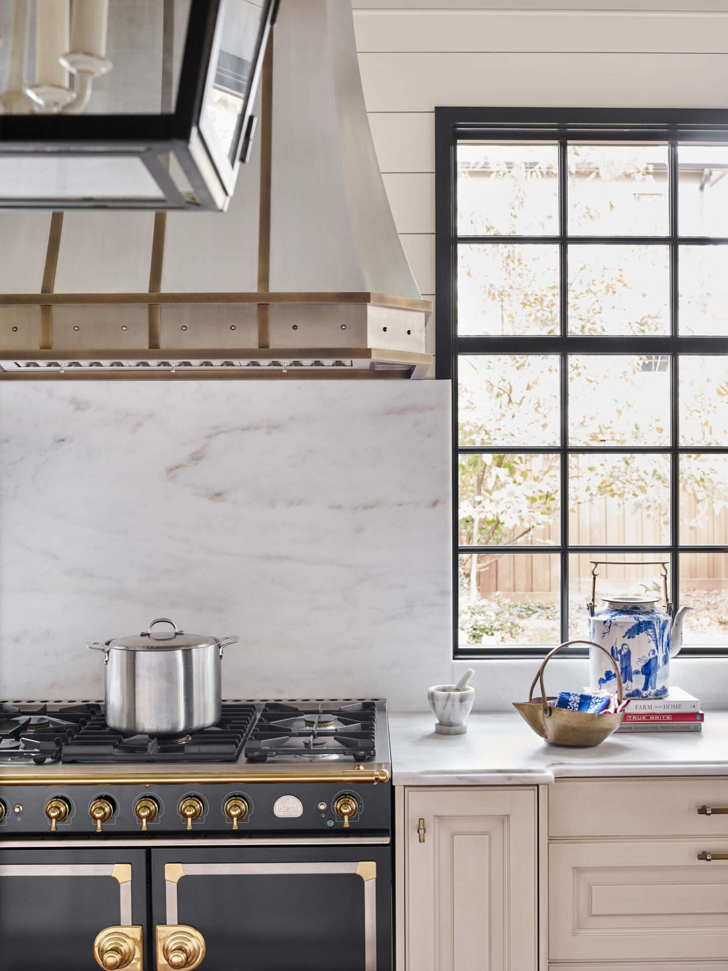 A modern kitchen features the Design Galleria black and gold stove with a silver pot, marble backsplash, cream cabinets, and a large window. On the counter are a blue and white jar, bowl, and small cup.