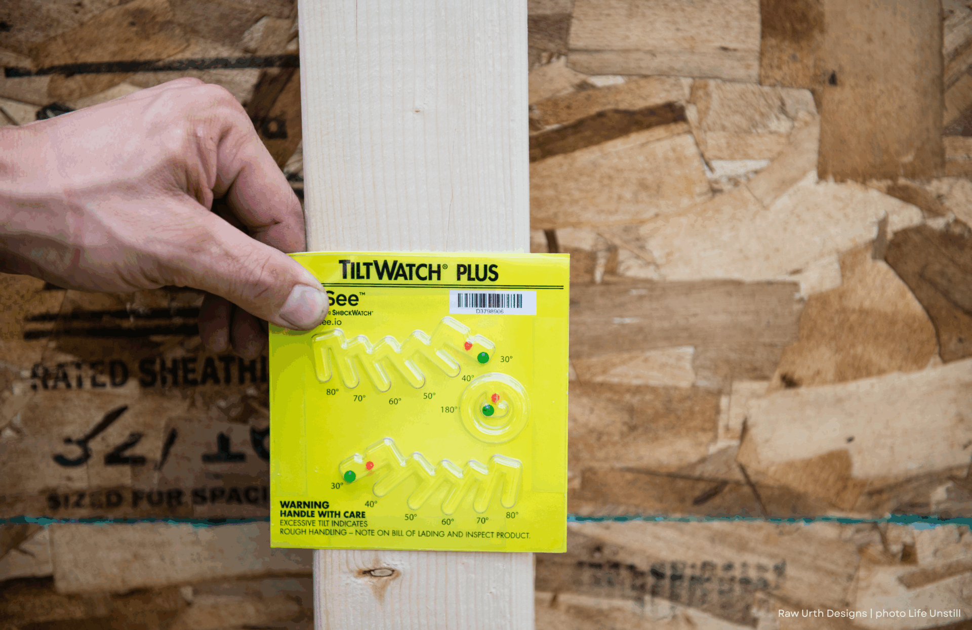 A hand holds a yellow 3 indicator against a wooden board, with an OSB plywood surface in the background. The 3 monitors a package's tilt during shipping or handling.