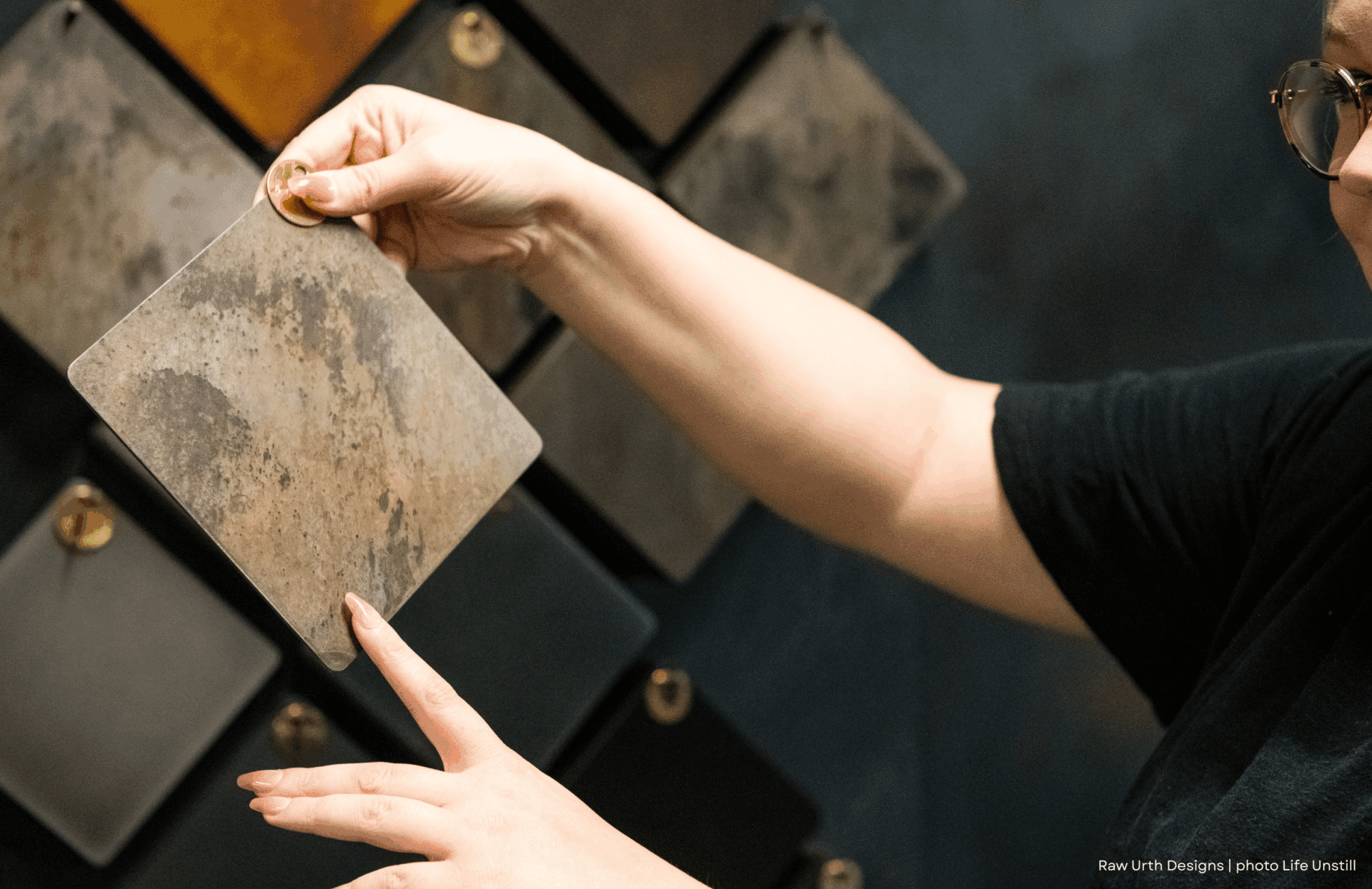 A person holds up a square sample of Product Name: 4 with a weathered texture, comparing it to similar samples displayed on a wall.