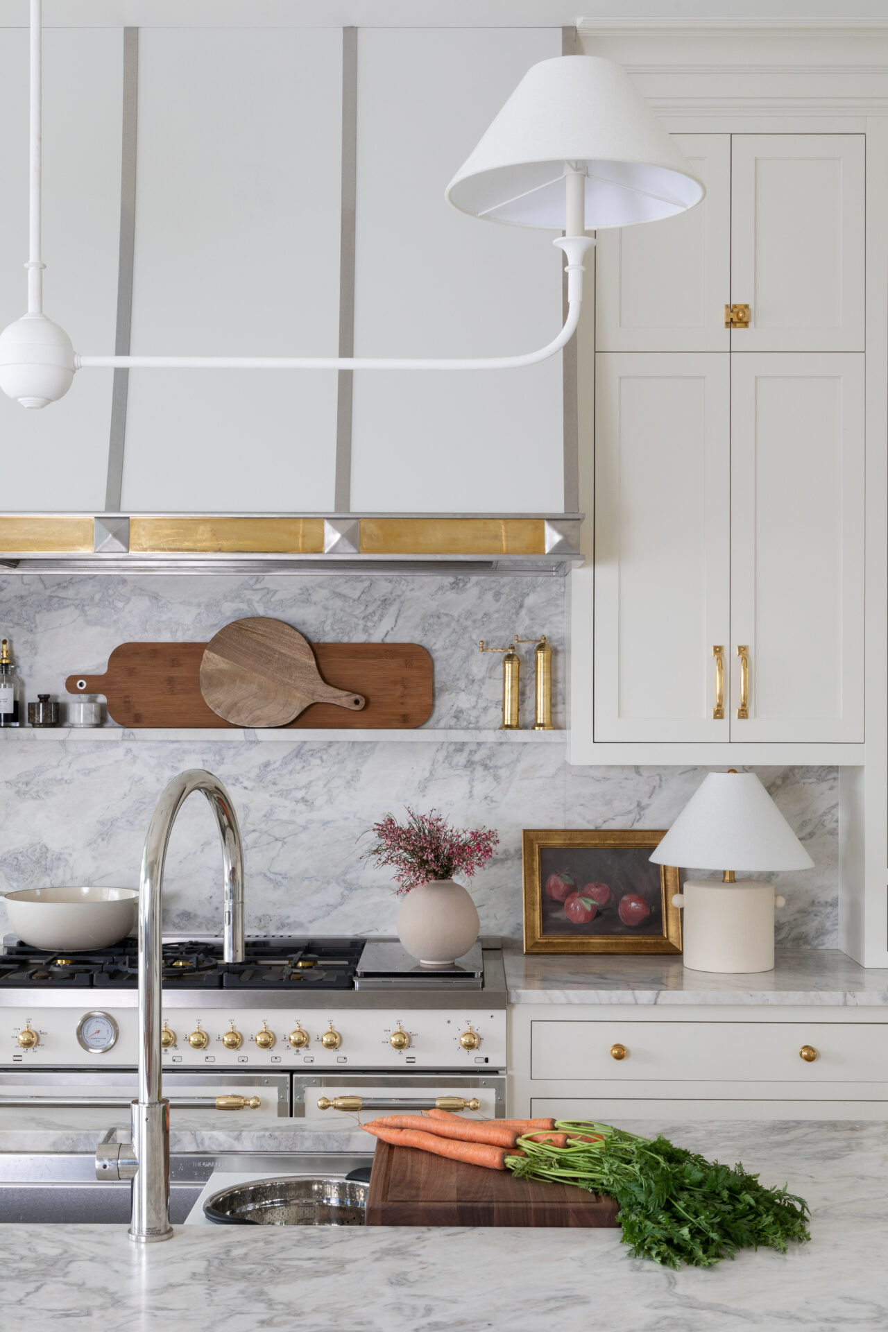 A bright, elegant kitchen featuring the 5354 Steel RH Enamel with SS Polished and Brass Polished faucet, marble countertops, gold-accented stovetop, cutting board with carrots, flowers in a vase, framed art, sleek lamp, and tall cabinets.