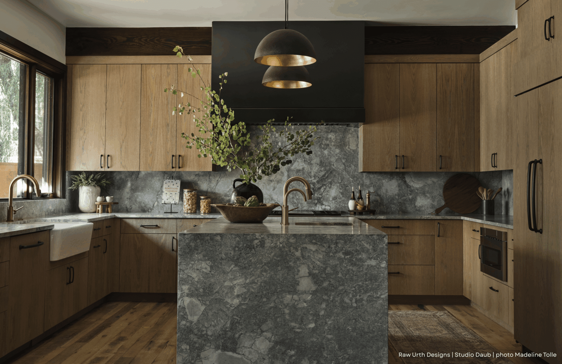 RU_Blog Images A bright kitchen featuring RU_Blog Images: light wood cabinets, gray marble surfaces, a large island with sink, black pendant lights, and greenery in a vase, bathed in sunlight from a nearby window.