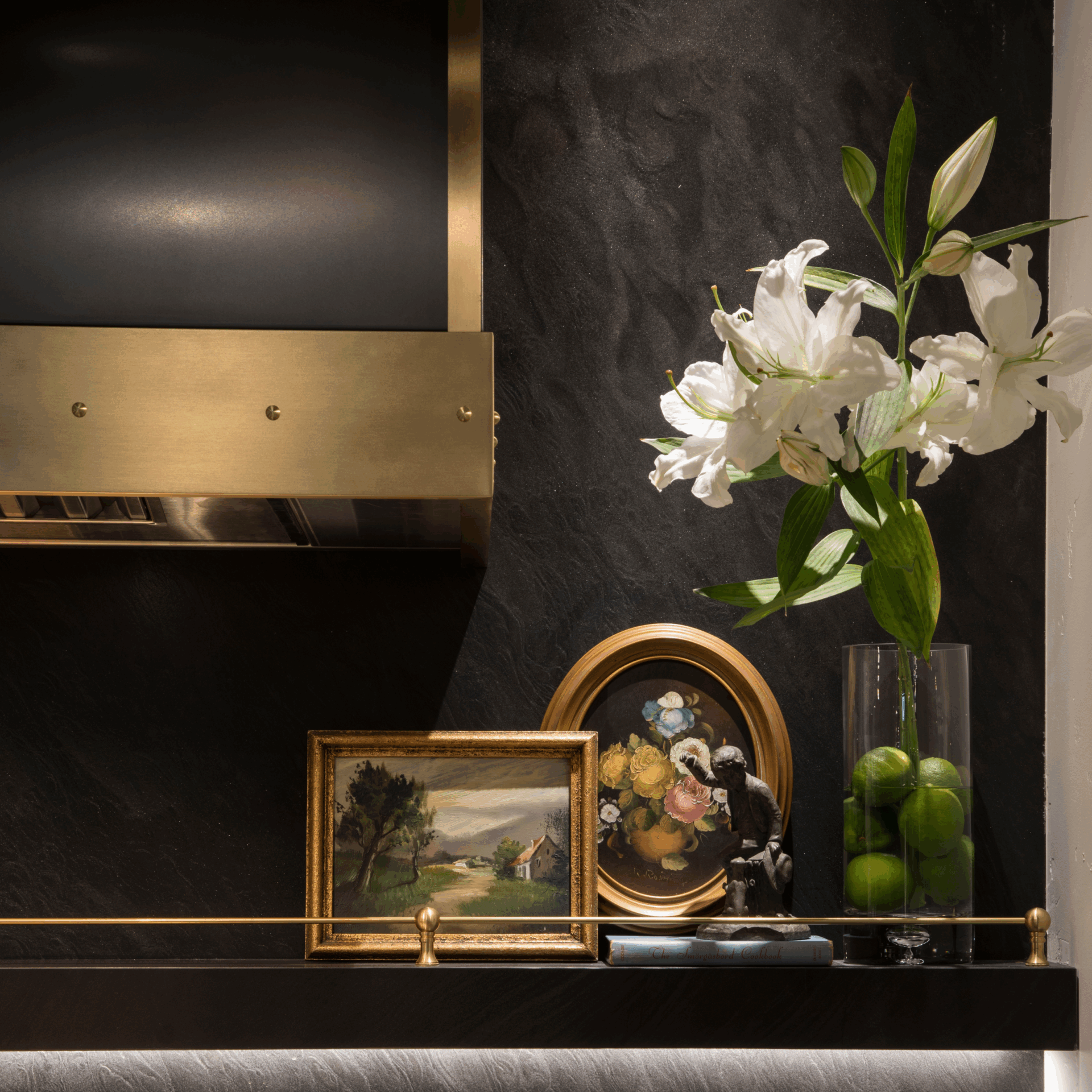 A Raw Urth Designs shelf from Cooper Pacific Kitchens displays a Meghan Beierle-O’Brien photo, an oval floral piece, a small figurine, and a tall vase with white lilies and green limes against a dark textured wall.