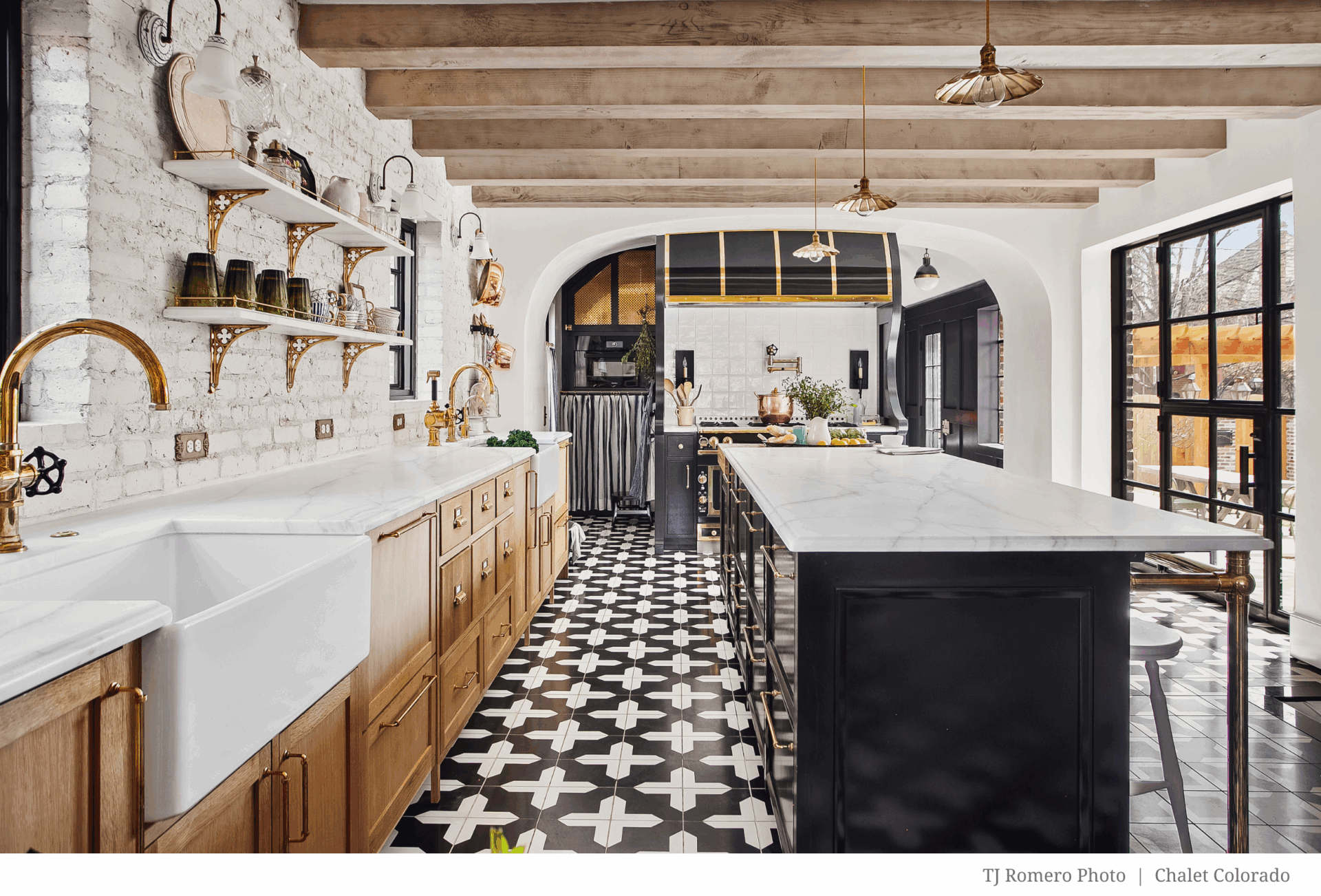 Image features wooden beams, white brick walls, black and white tile flooring, gold fixtures, open shelves, a farmhouse sink, marble countertops, and large windows for abundant natural light.