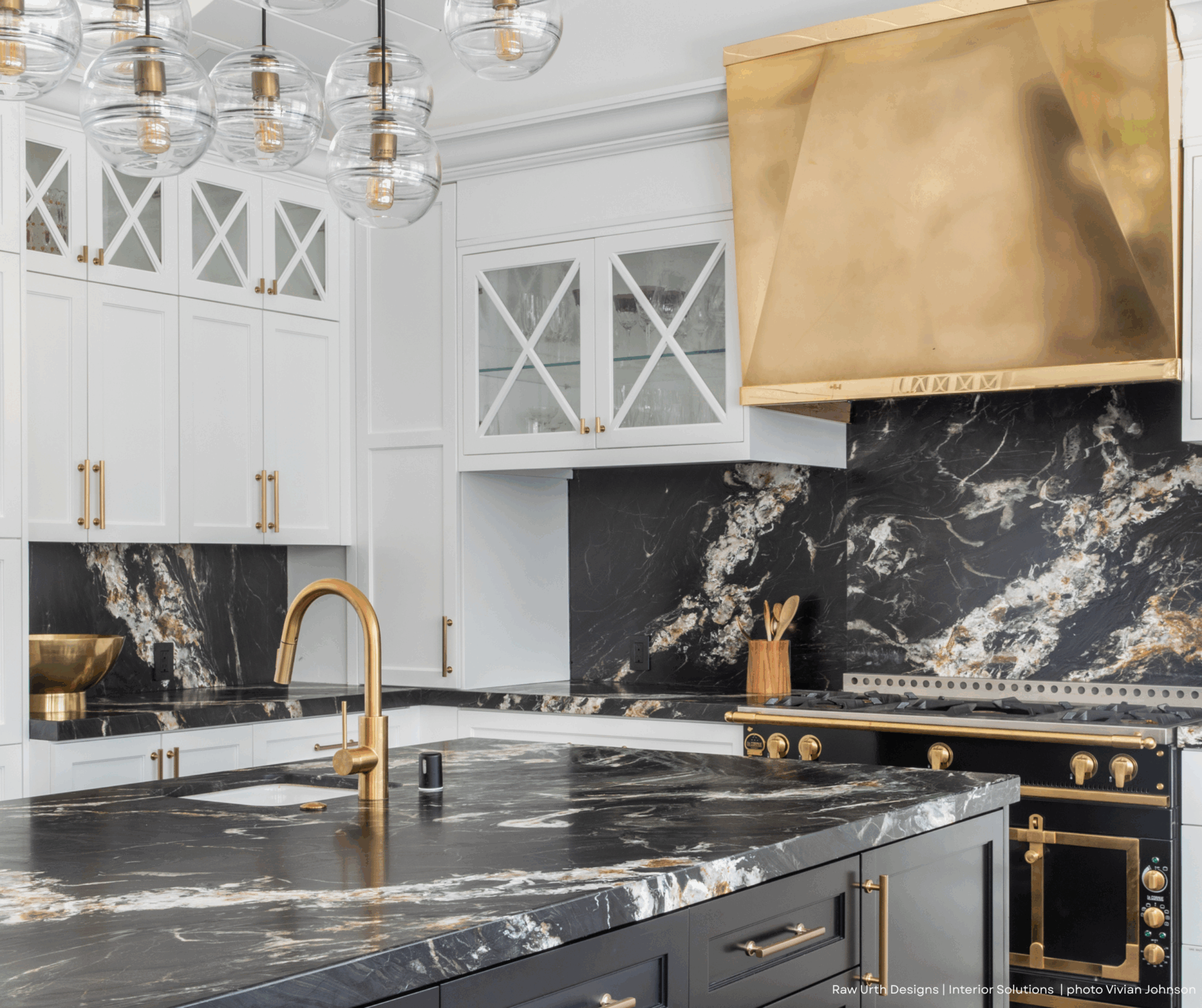 Modern kitchen with white cabinets, gold hardware, gold range hood, and black marble counters. Glass pendant lights hang above the island with a gold faucet and a built-in sink. Product shown: 2.