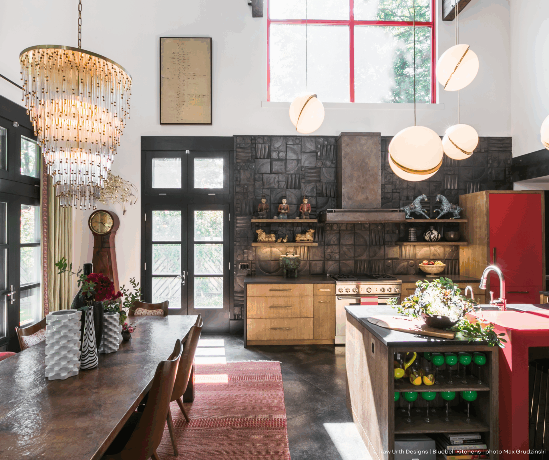 Bright kitchen with high ceilings, large windows, modern lighting, Cabinet color wooden cabinets, red fridge, black accents, and a dining table with flowers. Natural light floods the space.