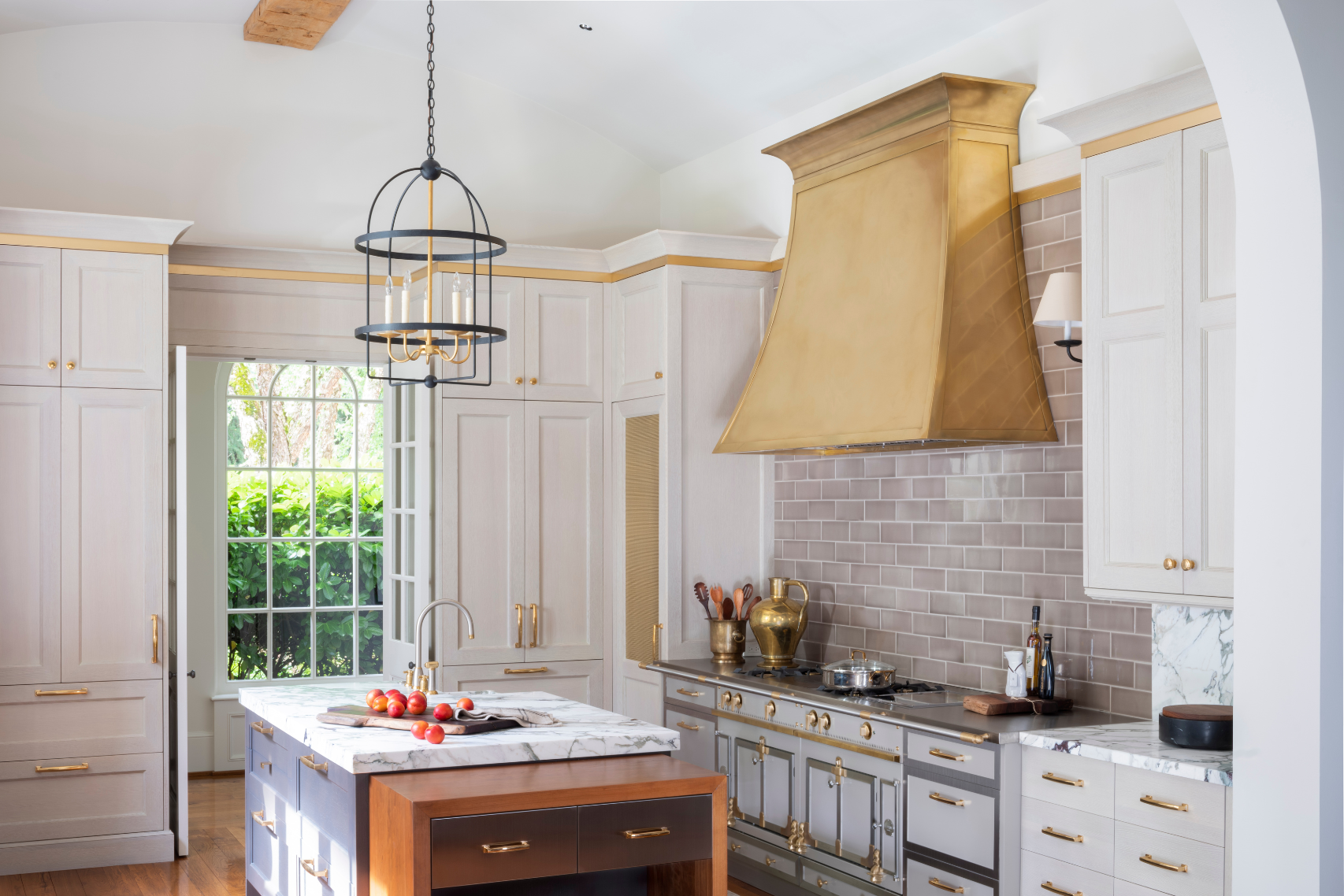A modern kitchen features 10’s light wood cabinets, a gold range hood, gray subway tile backsplash, marble countertops, a wood-base island, and pendant lights. Sunlight enters through large windows and a glass door.