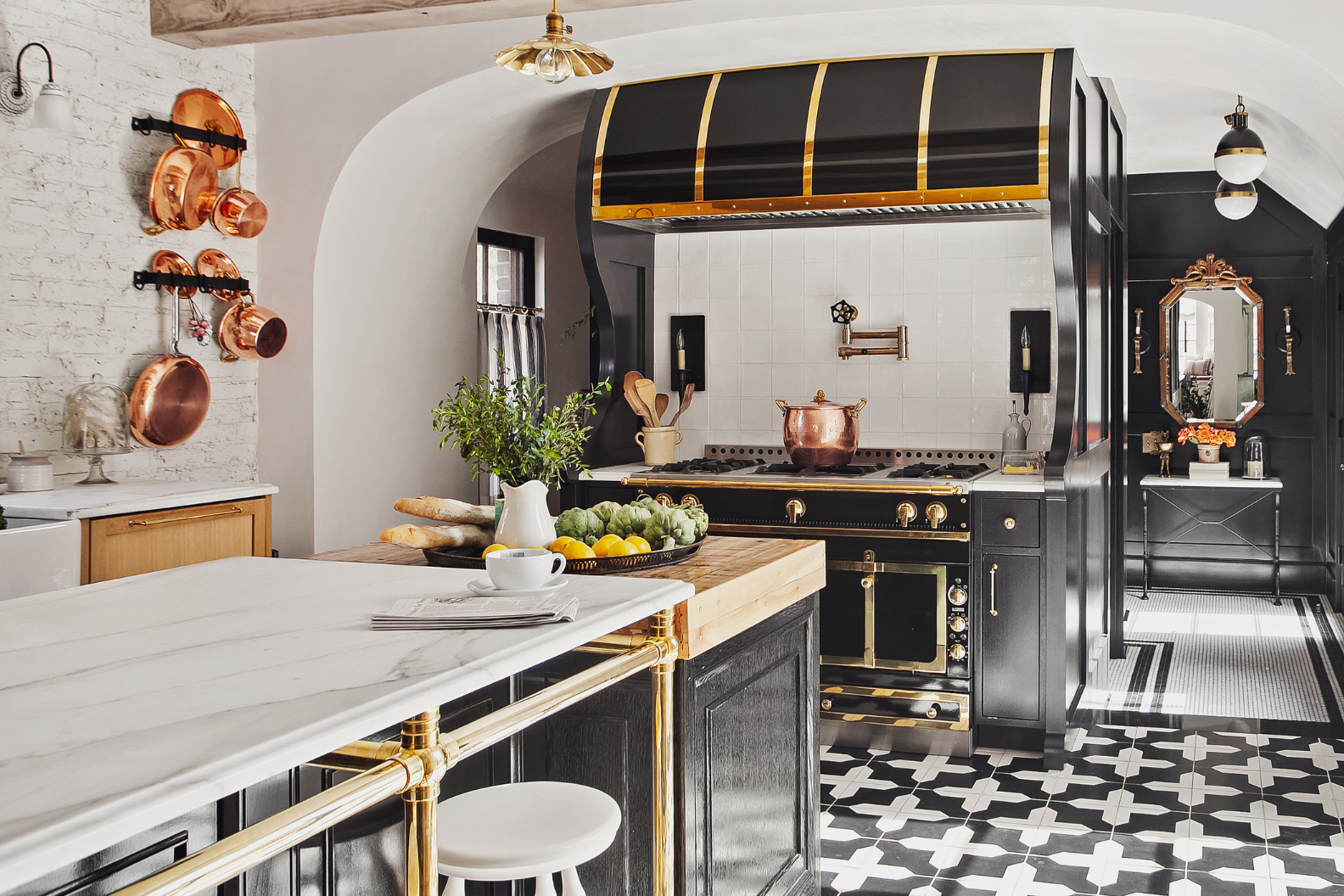 The 13 features a black and gold stove, white marble island, patterned tile floor, copper pots on the wall, and a blend of modern and vintage decor. Fresh bread, fruit, and greenery adorn the island.