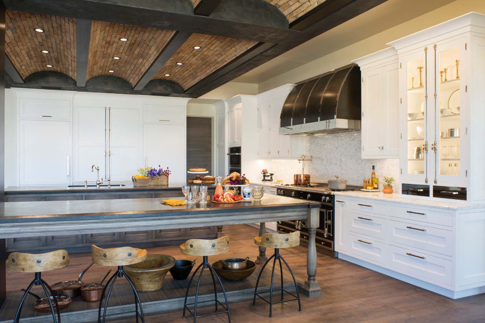 The 14 features a spacious modern kitchen with white cabinets, a large gray island with wooden stools, stainless steel appliances, exposed brick ceiling accents, and countertops adorned with decorative items and fresh produce.