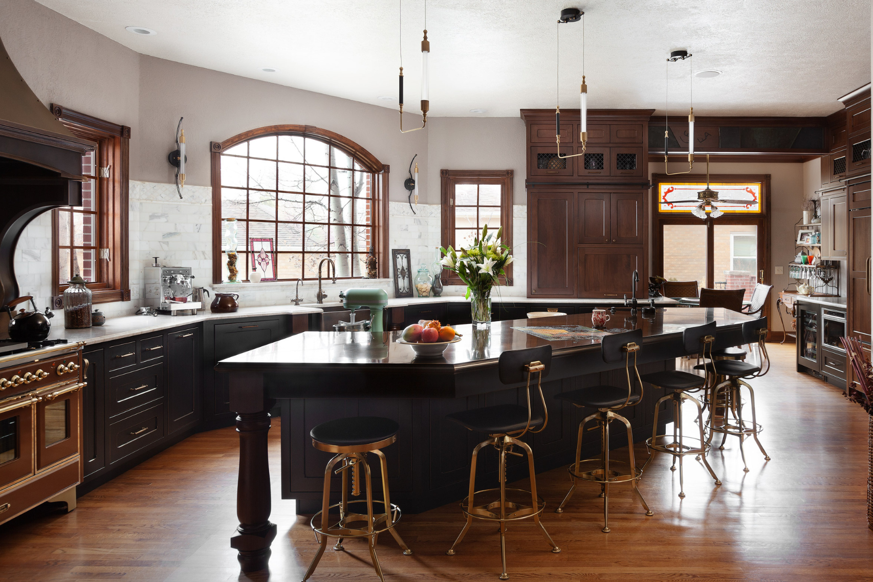 The 5 features dark wood cabinets, a large island with gold barstools, modern appliances, fresh flowers, arched windows for natural light, and warm hardwood floors that create an elegant kitchen space.